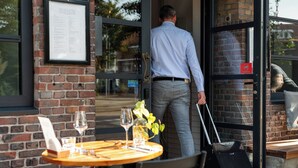 A man exiting a building with a suitcase, a menu on the wall, and a table with wine glasses and a plant. - Badhuis Hotel (Utrecht)