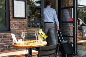 A man exiting a building with a suitcase, a menu on the wall, and a table with wine glasses and a plant.