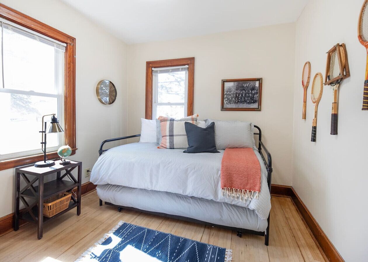 A bedroom with a bed, a nightstand, a window, a wall clock, and tennis rackets on the wall.