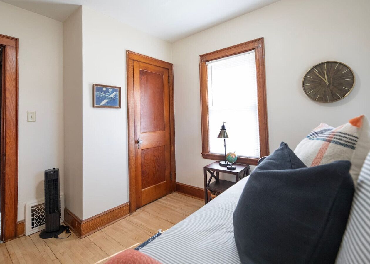 A room with a bed, a wooden door, a window, a wall clock, and a small table with a lamp and a globe.