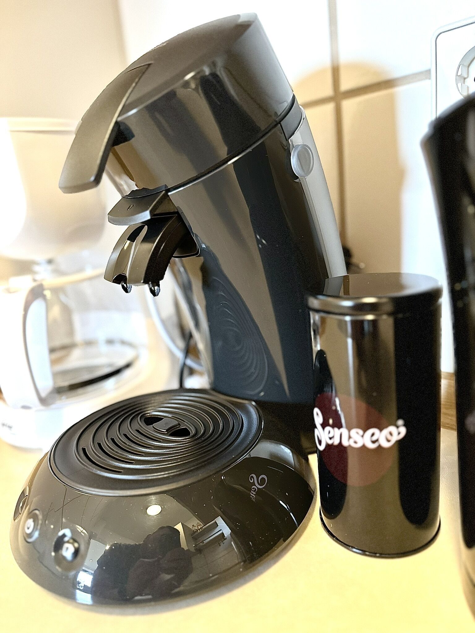 Une machine à café avec une base noire et un réservoir d’eau transparent, une tasse noire sur laquelle est inscrit le mot « Senseo  », et une cafetière blanche en arrière-plan.