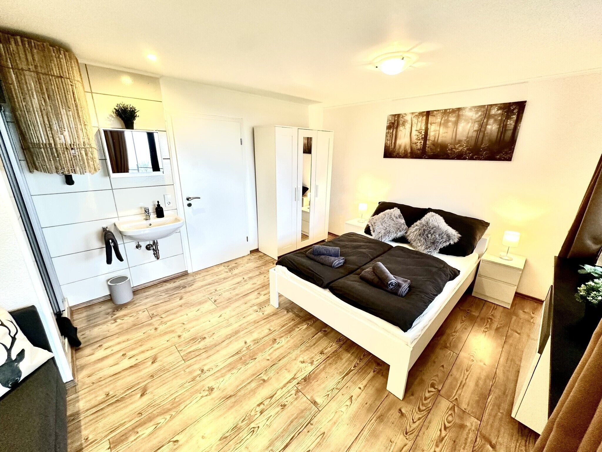Un dormitorio moderno con suelo de madera, una cama blanca con ropa de cama negra, una mesita de noche con lámpara y un espejo grande sobre el lavamanos.