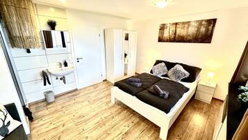 Un dormitorio moderno con suelo de madera, una cama blanca con ropa de cama negra, una mesita de noche con lámpara y un espejo grande sobre el lavamanos.