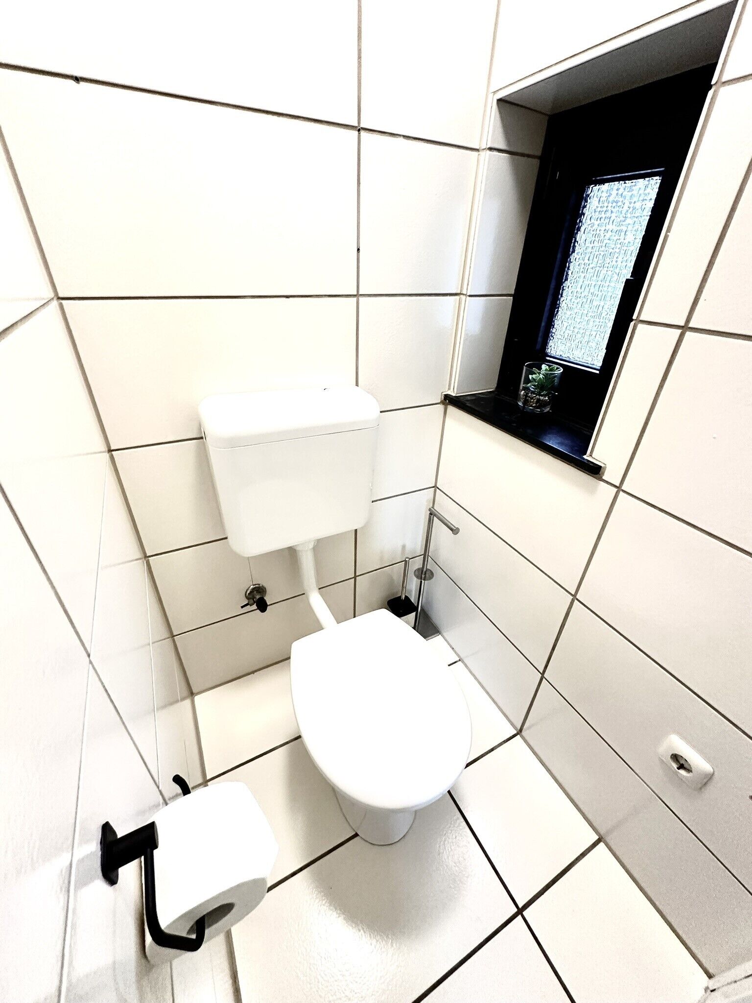 Pieni, moderni kylpyhuone, jossa on valkoiset laatat, wc-istuin, bidee ja ikkuna, josta on näkymä.