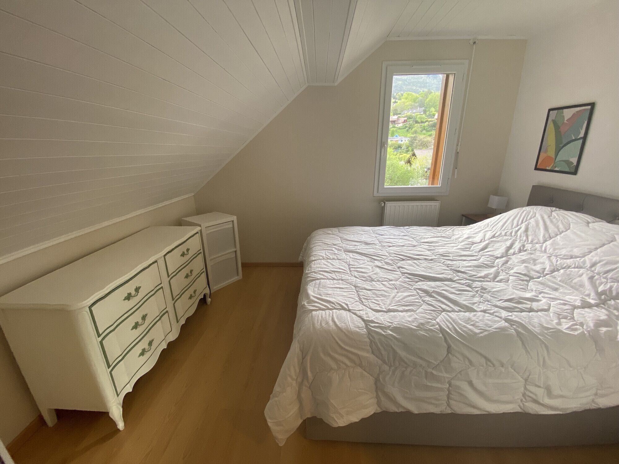 Un dormitorio con techo inclinado, cama con edredón blanco, cómoda con cajones, ventana con vista a follaje y un cuadro en la pared.