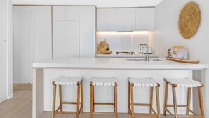 A modern kitchen with white cabinetry, a central island with stools, and a woven wall decoration. - Luxury Signature Stay Ocean Views 2BR Level 16 (Broadbeach)