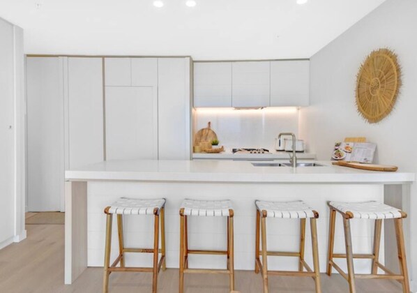 Een moderne keuken met witte kasten, een centraal eiland met krukken, en een wanddecoratie van geweven materiaal.