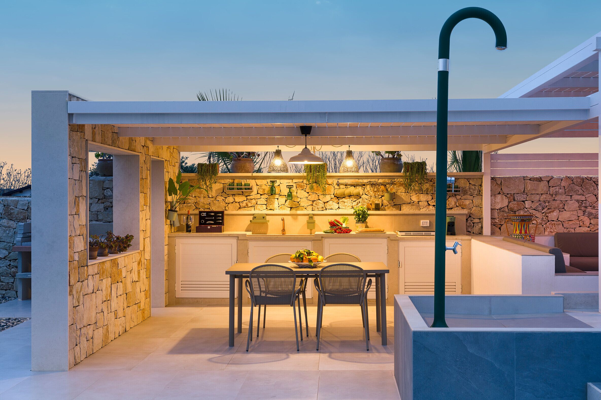 An outdoor kitchen area with stone walls, a dining table with chairs, and hanging plants.