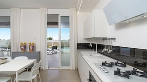 A modern kitchen with a large island, a built-in oven, and a gas stove. There is a dining area with a white table and chairs, and a view of an outdoor patio with a pool.