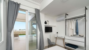 A modern hotel room with a sliding glass door to a balcony, a bed, a desk with a chair, and a built-in wardrobe with hanging clothes.