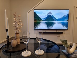 A room with a flat-screen TV displaying a mountain lake scene, two wine glasses on a glass table, a vase with dried flowers, and a candle. - Cloud Nine Elegant Escape Awaits (Christchurch)