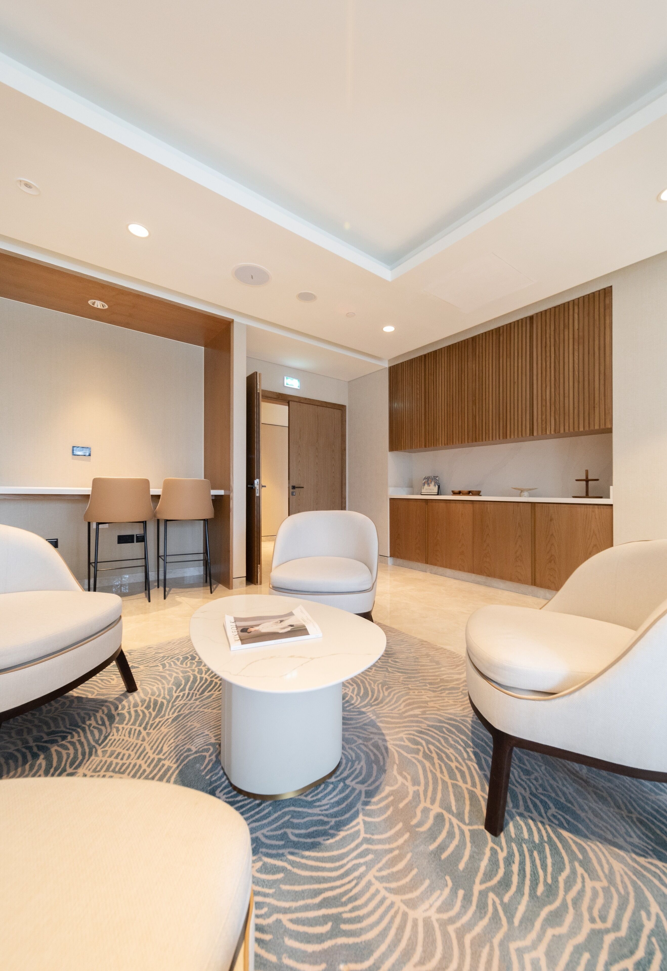 Ein modernes Hotelzimmer mit einer Sitzecke, bestehend aus zwei Stühlen und einem kleinen Tisch, einer Küchenzeile mit Holzschränken sowie einem gemusterten Teppich.