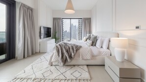 A modern bedroom with a large bed, a TV, a bedside lamp, and a view of the city through floor-to-ceiling windows. - EDEN'S Homes & Villas - Act One Act Two (Dubai)