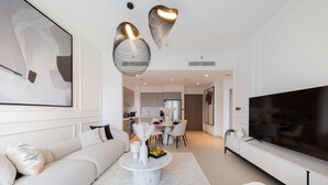 A modern living room with a large flat-screen TV, a white sofa, a coffee table, and a dining area in the background. - EDEN'S Homes & Villas - Act One Act Two (Dubai)