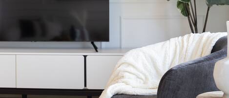 Un salon moderne avec un téléviseur à écran plat fixé à un mur blanc, un canapé gris foncé agrémenté d’un plaid blanc et une plante en pot aux feuilles vertes.