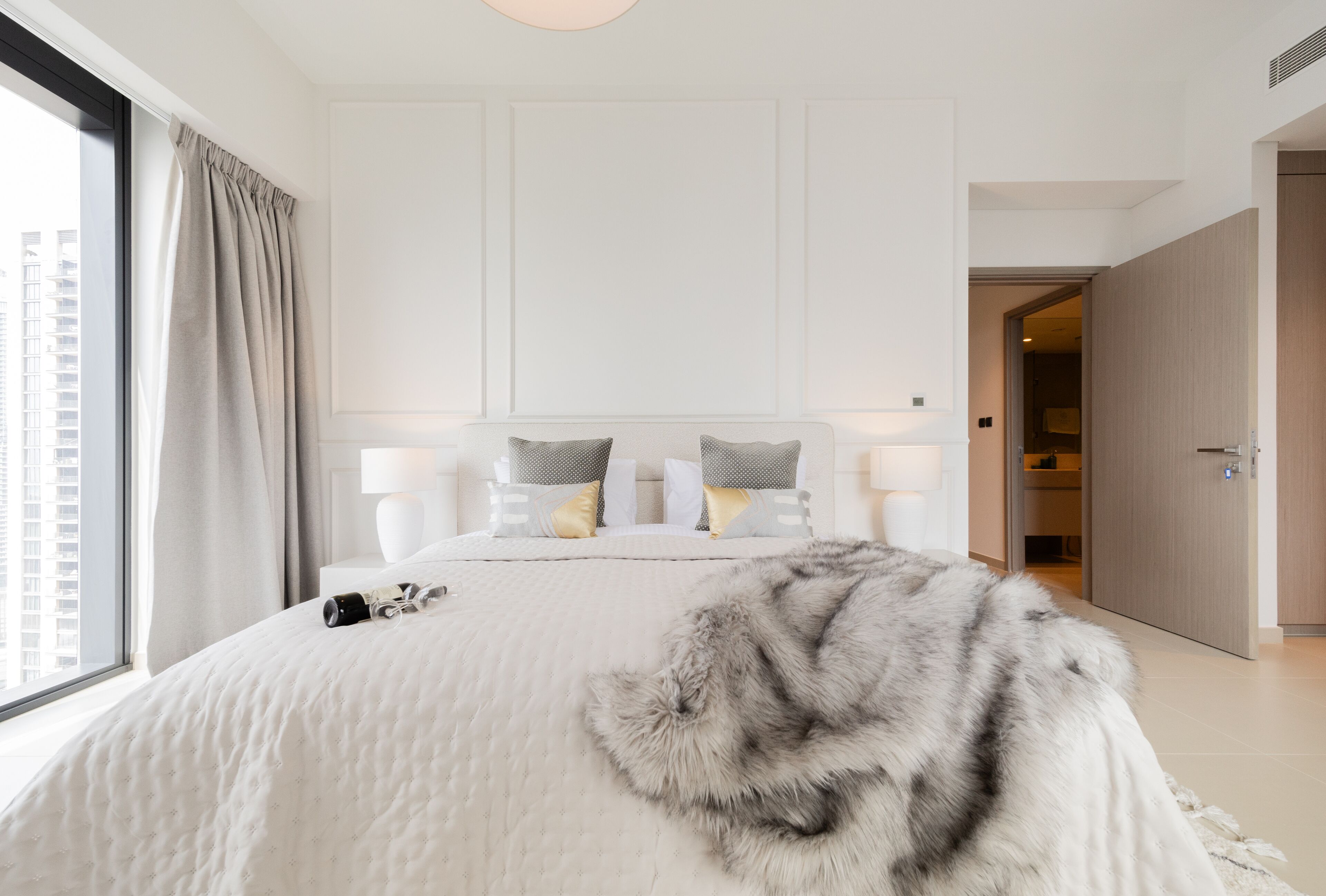 A modern hotel bedroom with a large bed, two bedside lamps, a fluffy throw blanket, and a view of the city through a large window.