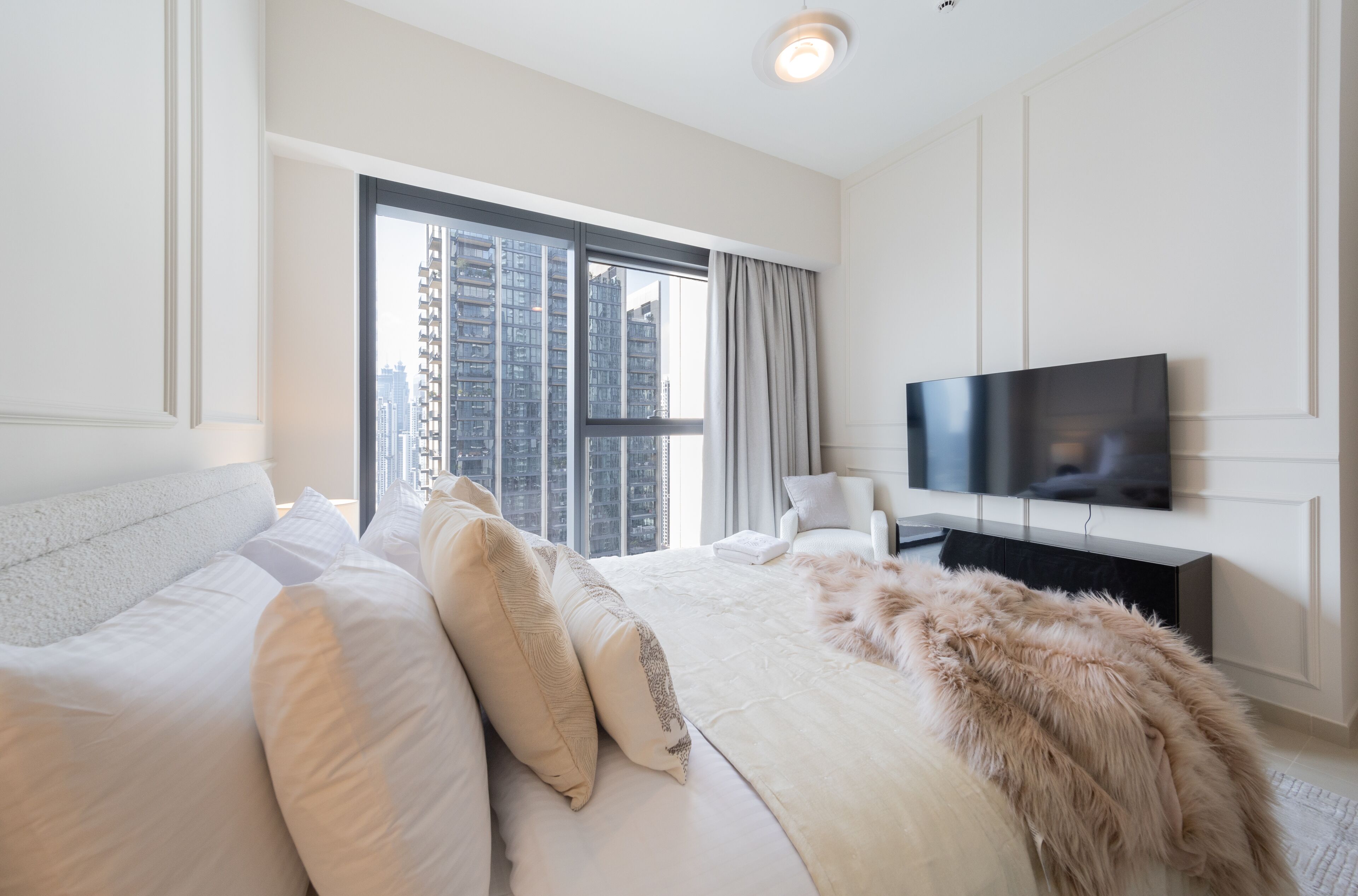 A modern hotel room with a large bed, a flat-screen TV, and a view of high-rise buildings through a large window.