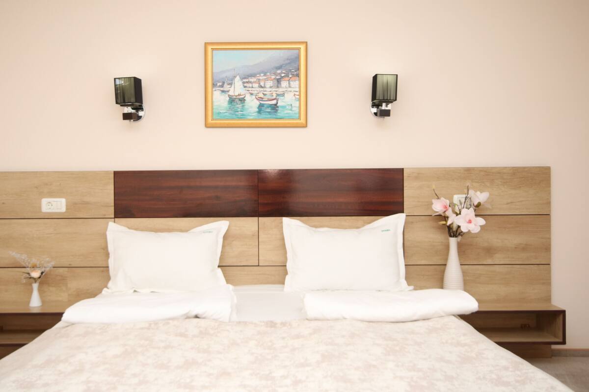 Une chambre d’hôtel avec une tête de lit en bois, deux oreillers blancs, un lit recouvert d’une couverture de couleur claire, un tableau représentant un paysage côtier et un vase de fleurs posé sur la table de chevet.
