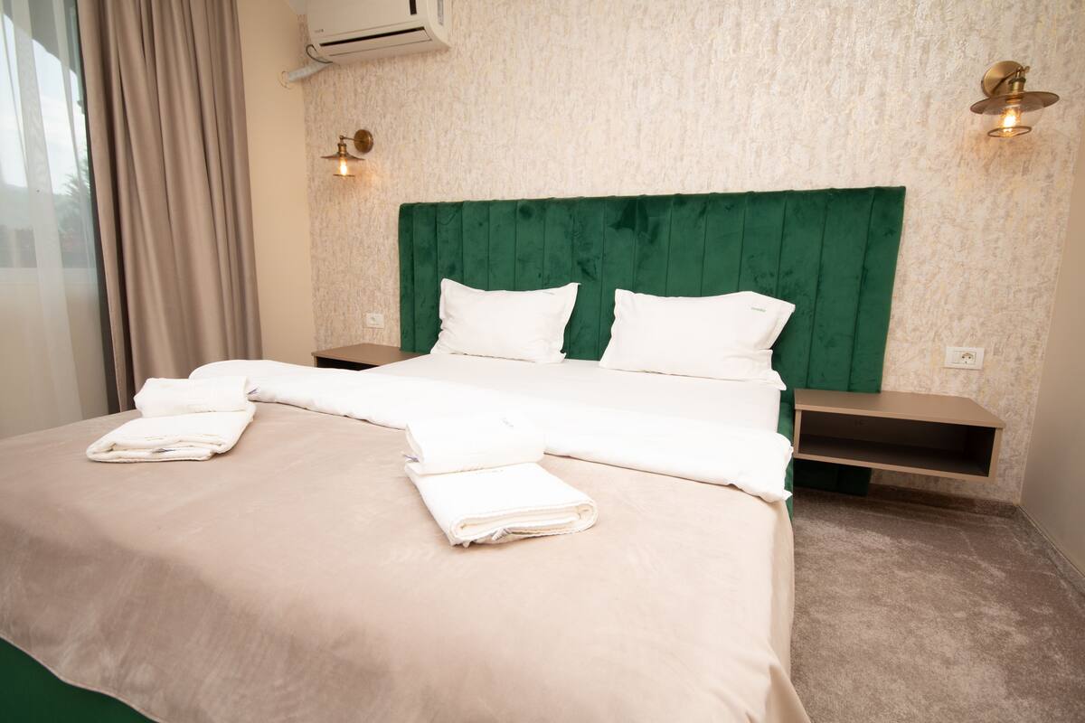 Un lit bien fait, avec du linge de lit blanc et des oreillers, une tête de lit recouverte d’un tissu vert et des tables de chevet avec des lampes.