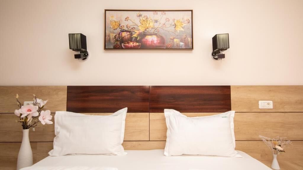 A hotel room with a wooden headboard, two white pillows, a vase with flowers, and a painting above the bed.