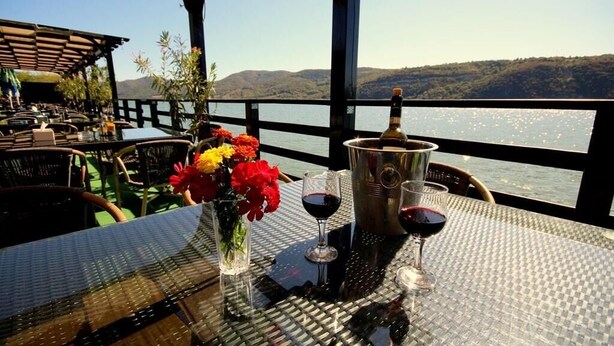 Ein Tisch mit Champagnerflasche, zwei Weingläsern und einer Blumenvase auf einer Terrasse mit Blick auf ein Gewässer und Hügel.
