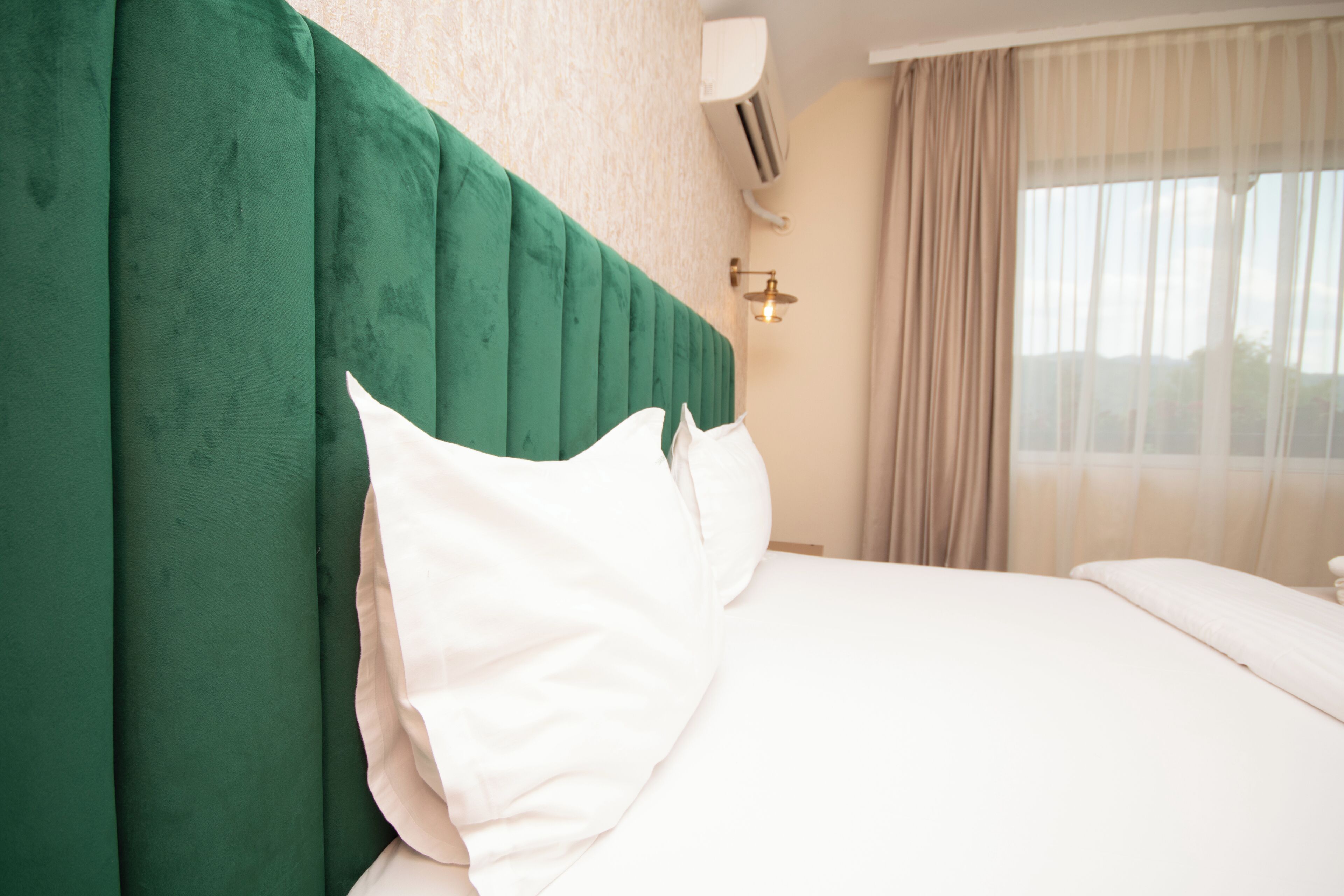 Een hotelkamer met een groen hoofdeinde, witte kussens, een beige muur en een raam met gordijnen.