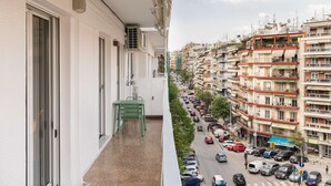 Un balcón con una silla verde, una ventana y vista a una calle concurrida con coches estacionados y edificios de varios pisos.