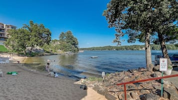 Una playa con un cartel que indica prohibido el acceso de mascotas, orilla arenosa y un lago tranquilo al fondo.