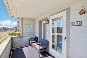 A covered porch with two chairs and a table, a door with a number sign, and a view of the street.