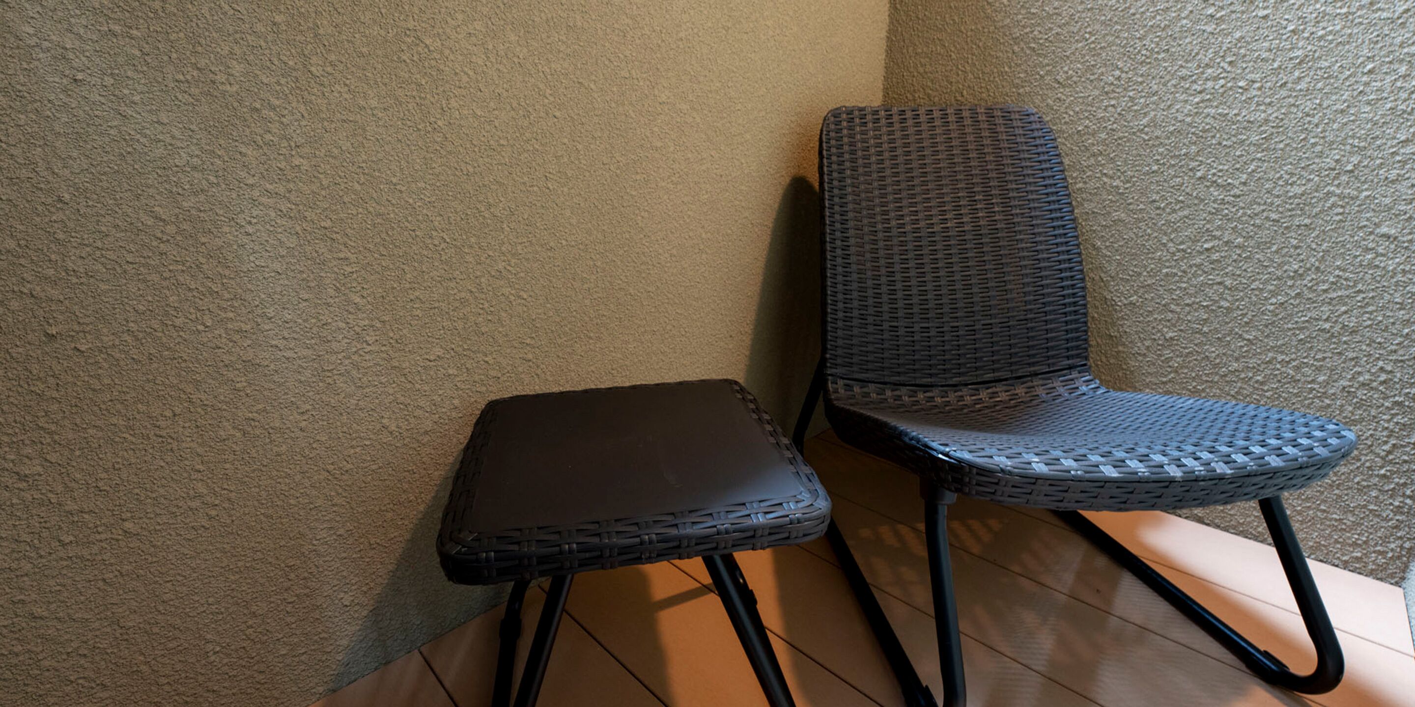A corner with a wicker chair and a small table, both with black metal legs, against a textured beige wall.