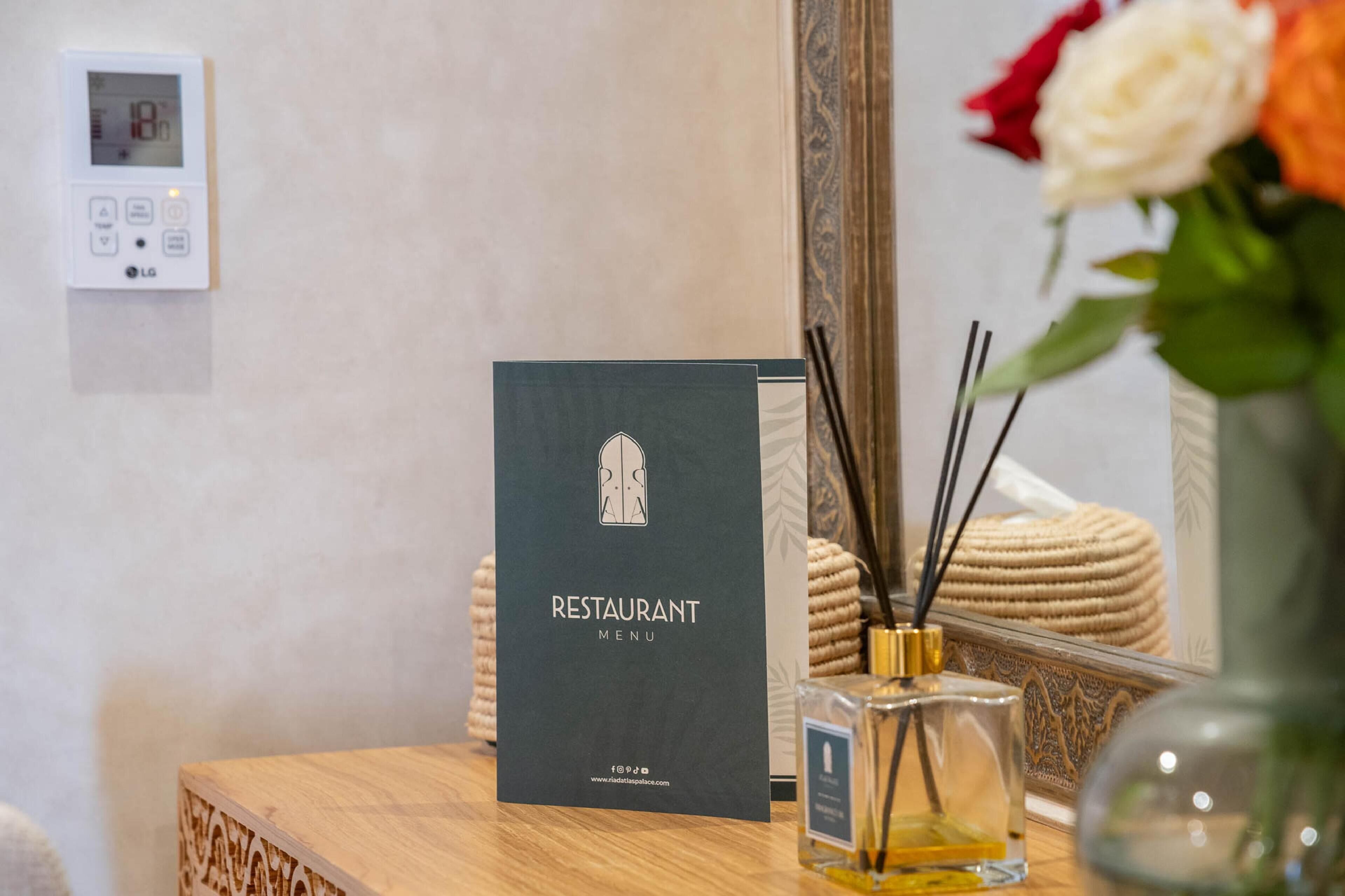 A restaurant menu sign on a wooden table with a mirror reflecting a vase of flowers and a bottle of perfume.