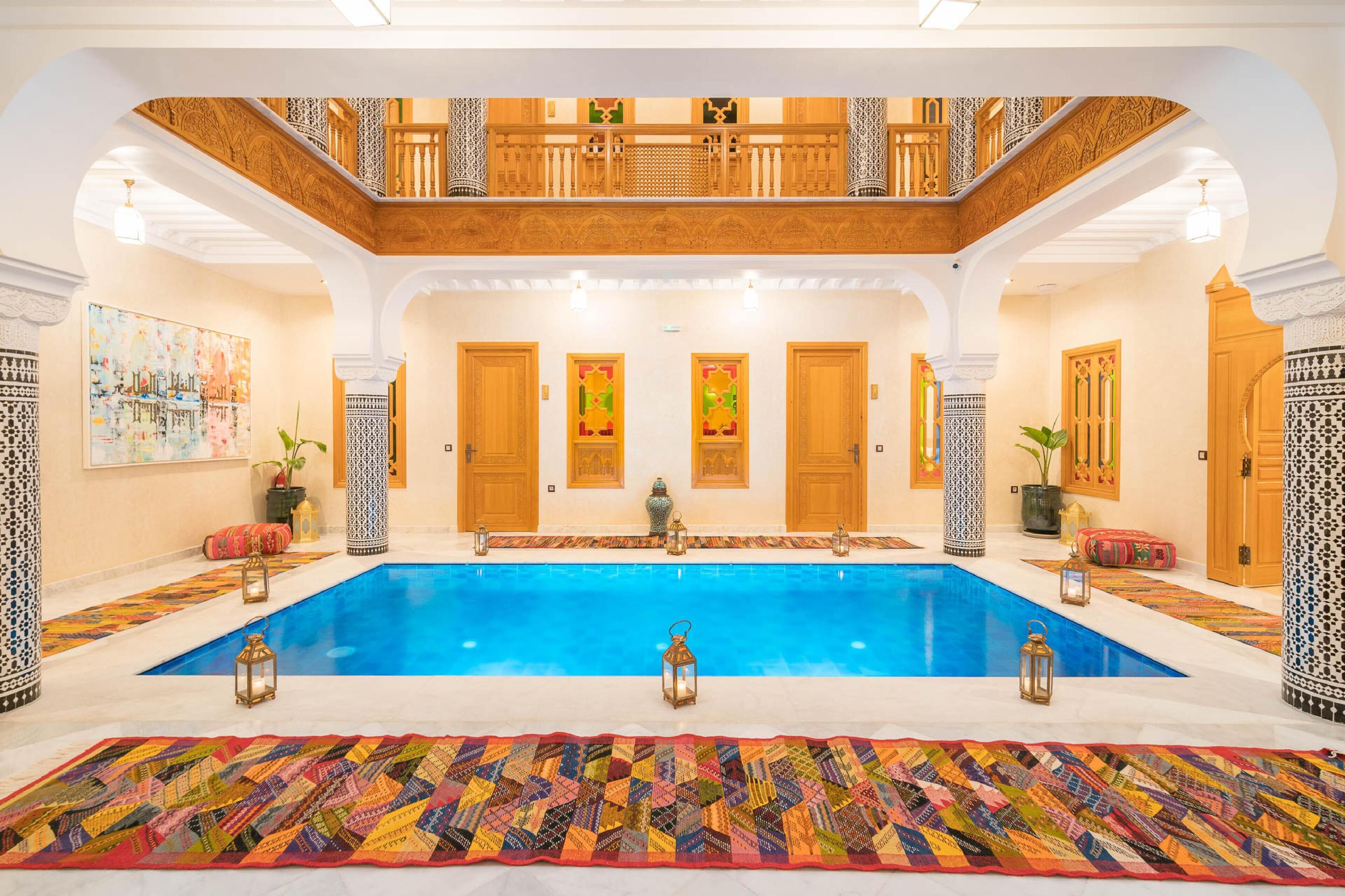 A spacious indoor swimming pool with a wooden balcony, ornate doors, and patterned rugs.