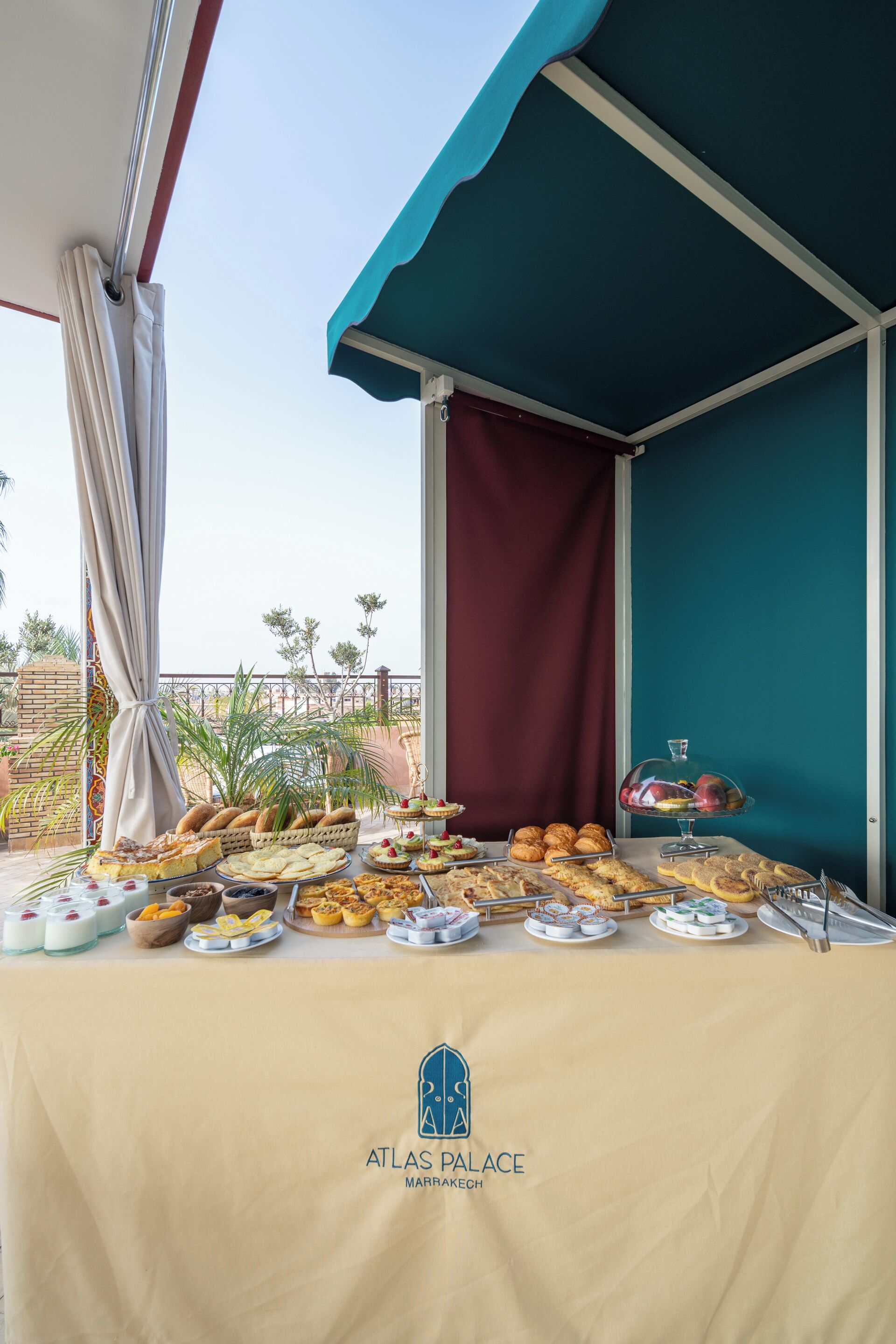 A buffet table with various food items and a glass display case, set up under a blue awning with a view of palm trees and a building in the background.