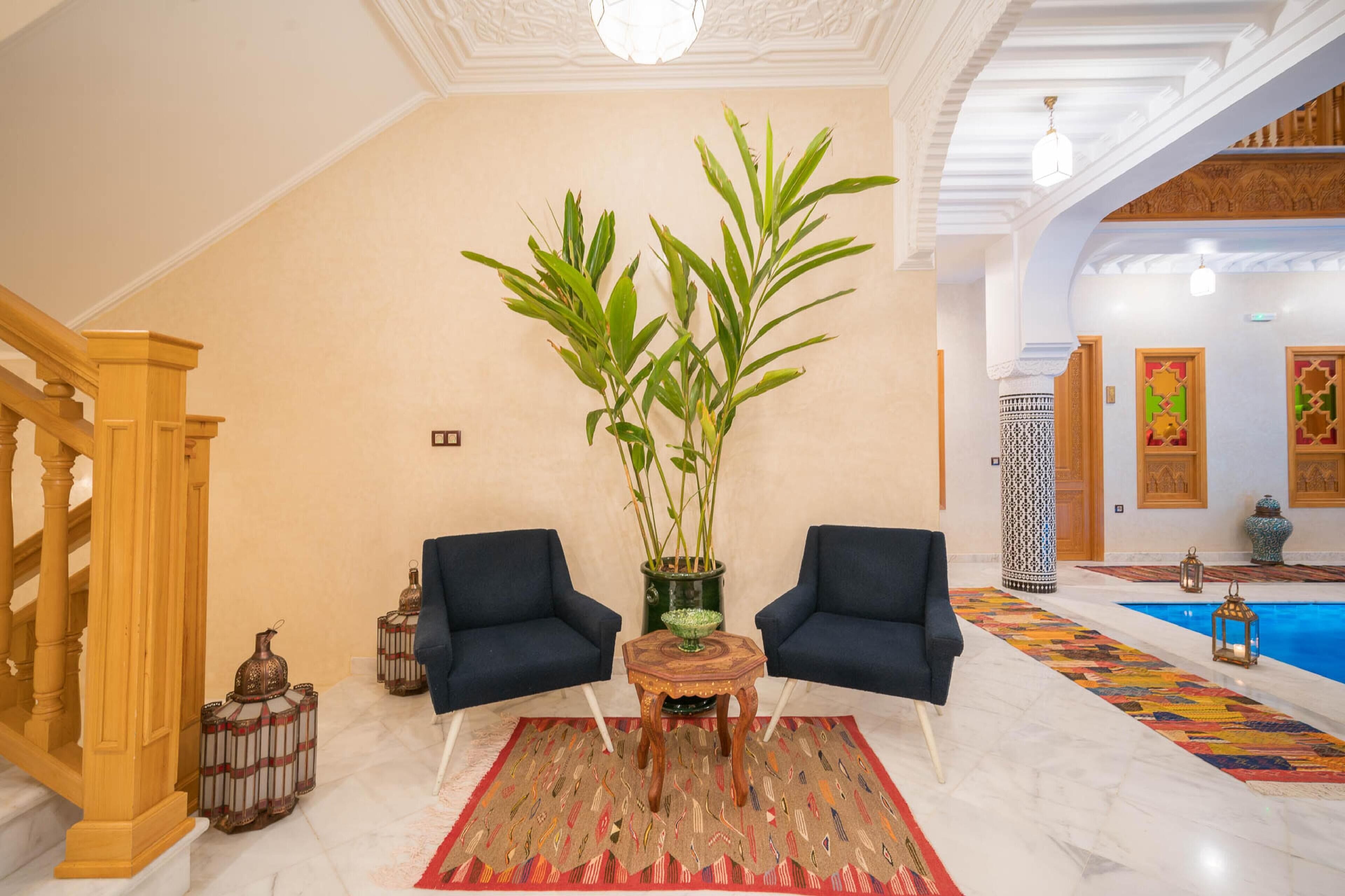 A spacious lobby with a marble floor, two dark blue armchairs, a large bamboo plant, and a colorful rug.