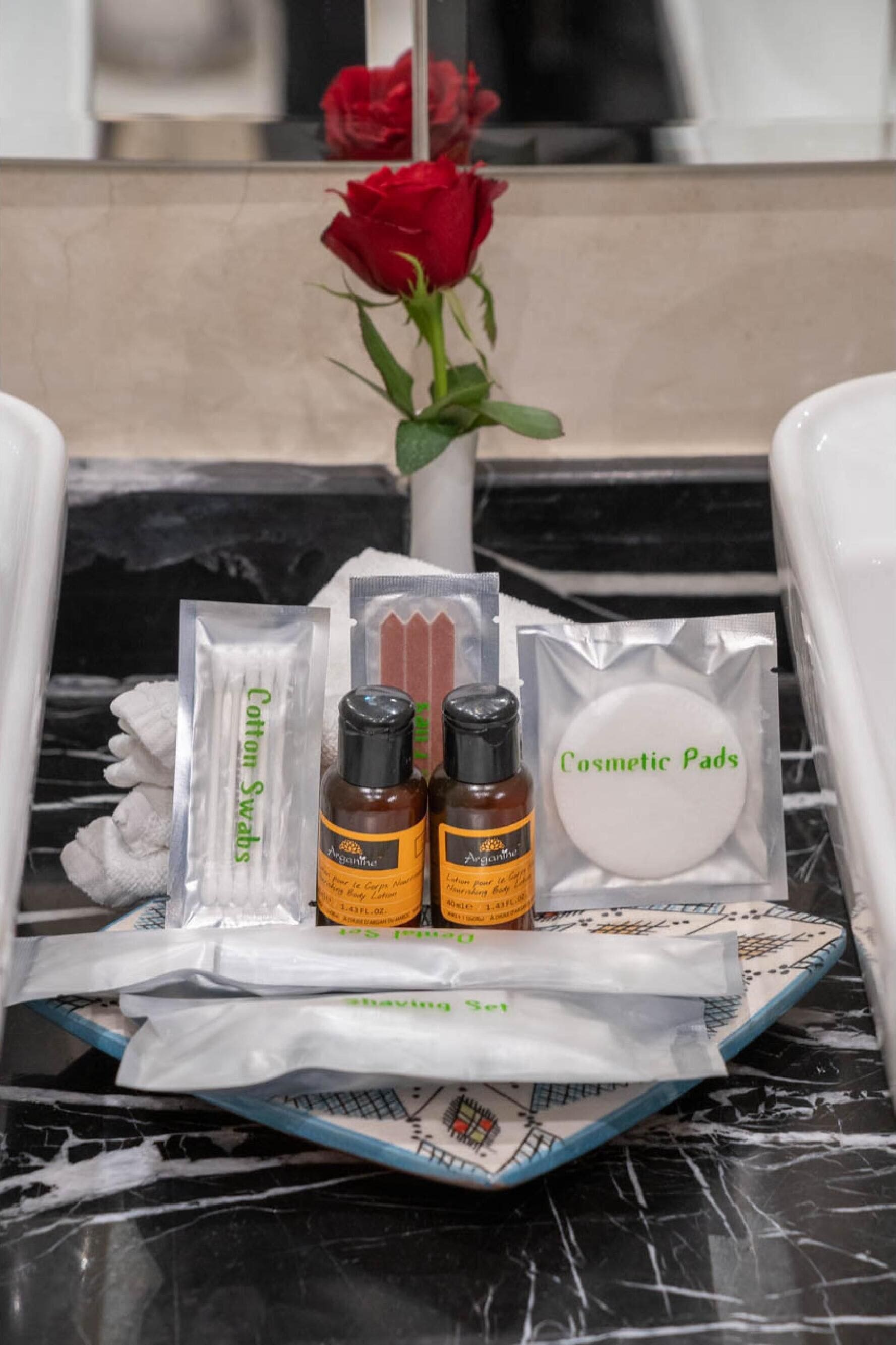 A tray with cosmetic items including cotton swabs, essential oils, and cosmetic pads, placed on a marble surface with a red rose in the background.