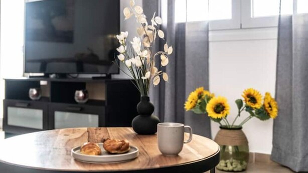 Een ronde houten tafel met een bord croissants, een grijze mok en een vaas met zonnebloemen.