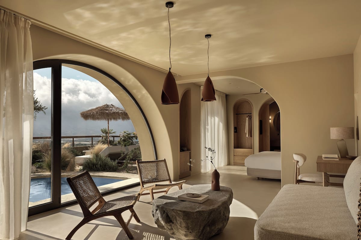 Une chambre d’hôtel moderne dotée d’une grande fenêtre en arc d’où l’on peut admirer une piscine et une vue sur l’océan.
