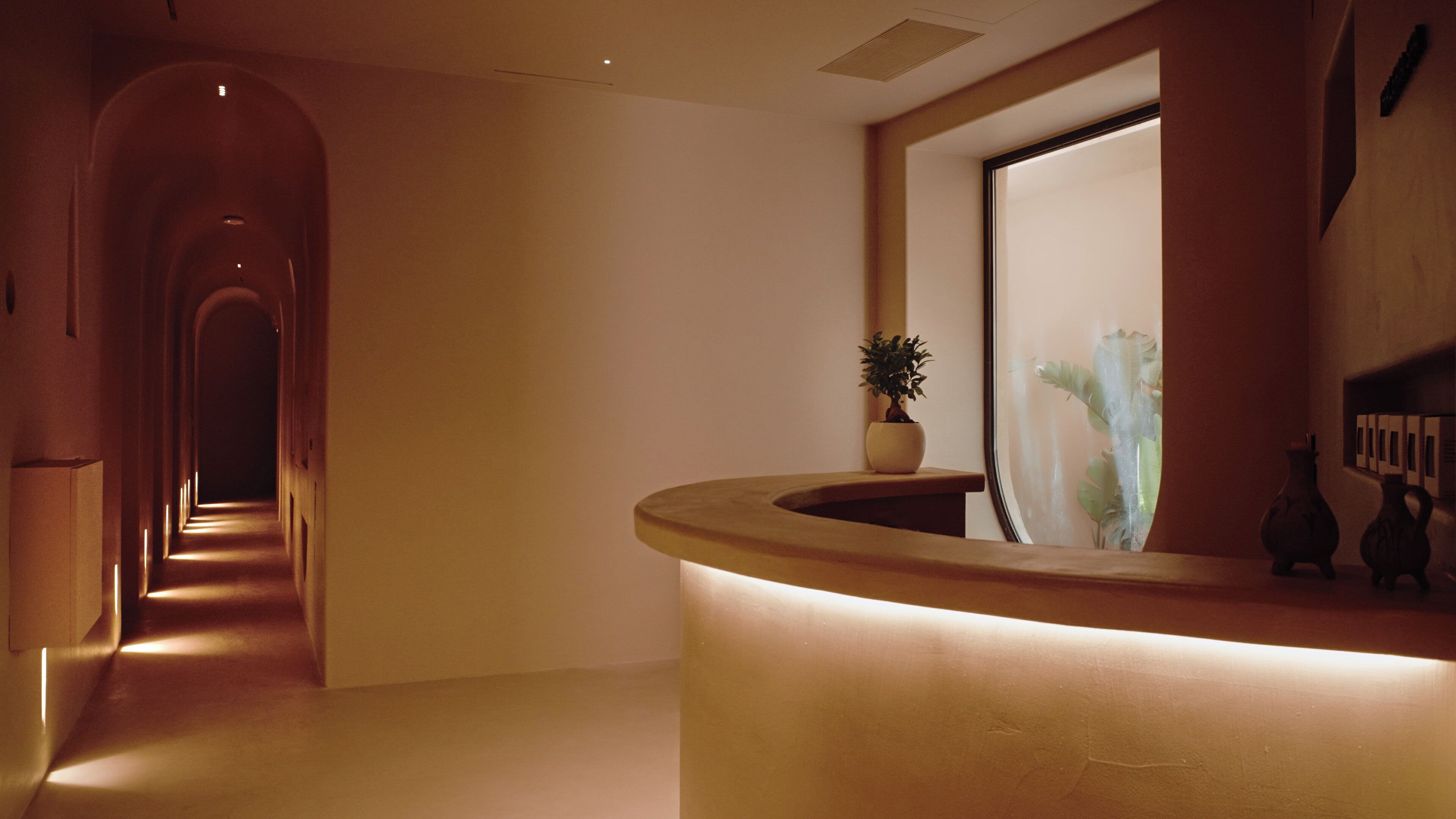 A hotel reception area with a curved reception desk, a potted plant, and a long hallway leading to an arched doorway.