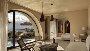Ein geräumiger Raum mit großem Bogenfenster mit Blick auf Pool und Meer, eine gemütliche Sitzecke mit Stühlen und einem Steintisch sowie ein separater Schlafbereich mit Bett und Nachttisch.