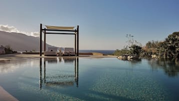 A clear pool with a wooden structure, lounge chairs, and a view of the ocean.
