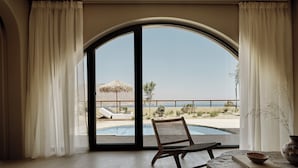Ein Zimmer mit einem großen Bogenfenster, einem Korbstuhl, einem Steintisch und Blick auf einen Pool und eine Außenanlage.