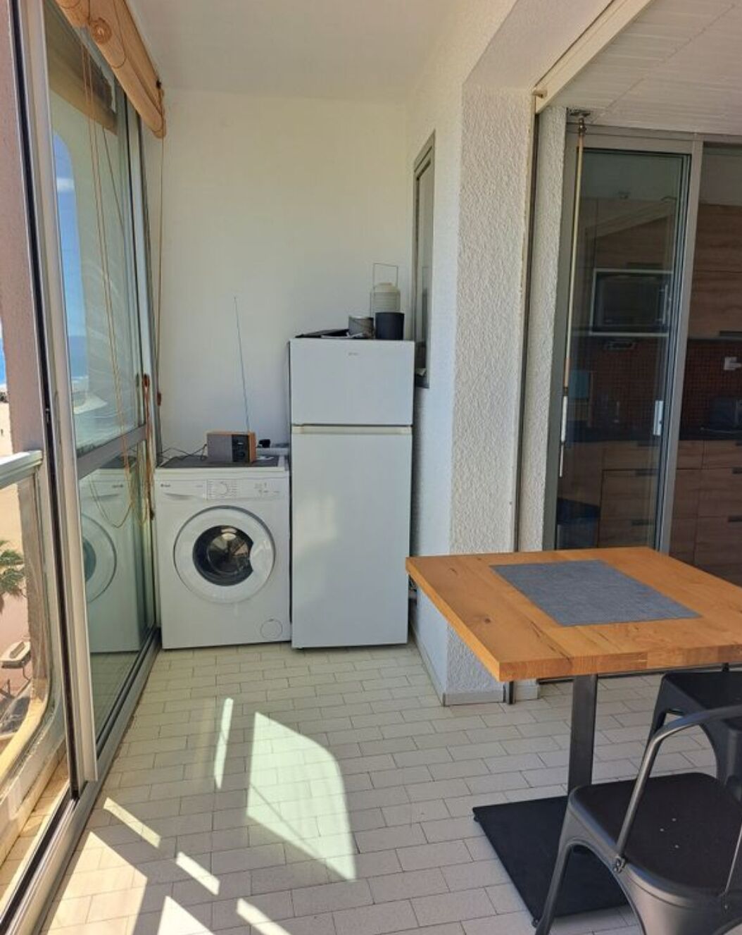 A small laundry area with a washing machine and a refrigerator, a wooden table with a chair, and a sliding glass door leading to a balcony.