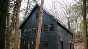 Une cabane en bois sombre, dotée de nombreuses fenêtres et d’une cheminée, entourée d’une forêt.