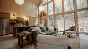Un vaste espace de vie avec une grande fenêtre, un canapé vert, une table basse en bois et une cuisine dotée de meubles en bois et d’un réfrigérateur.