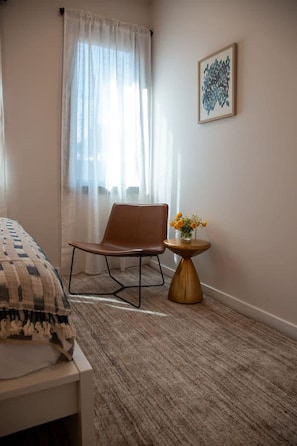 Un dormitorio con una cama, una silla, una mesita redonda con un florero y una ventana con cortinas de gasa.