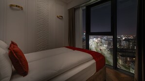 Een slaapkamer met een groot raam, een bed met witte beddengoed en een rode kussen, en uitzicht op een stadsbeeld 's nachts.