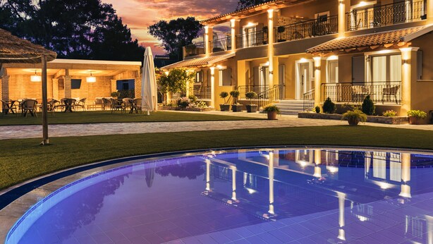 A poolside view of a hotel with a large pool, outdoor seating area, and a well-lit building with multiple balconies. - Comissa Luxury Suites (Corfu)