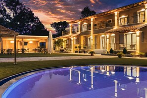 A poolside view of a hotel with a large pool, outdoor seating area, and a well-lit building with multiple balconies. - Comissa Luxury Suites (Corfu)