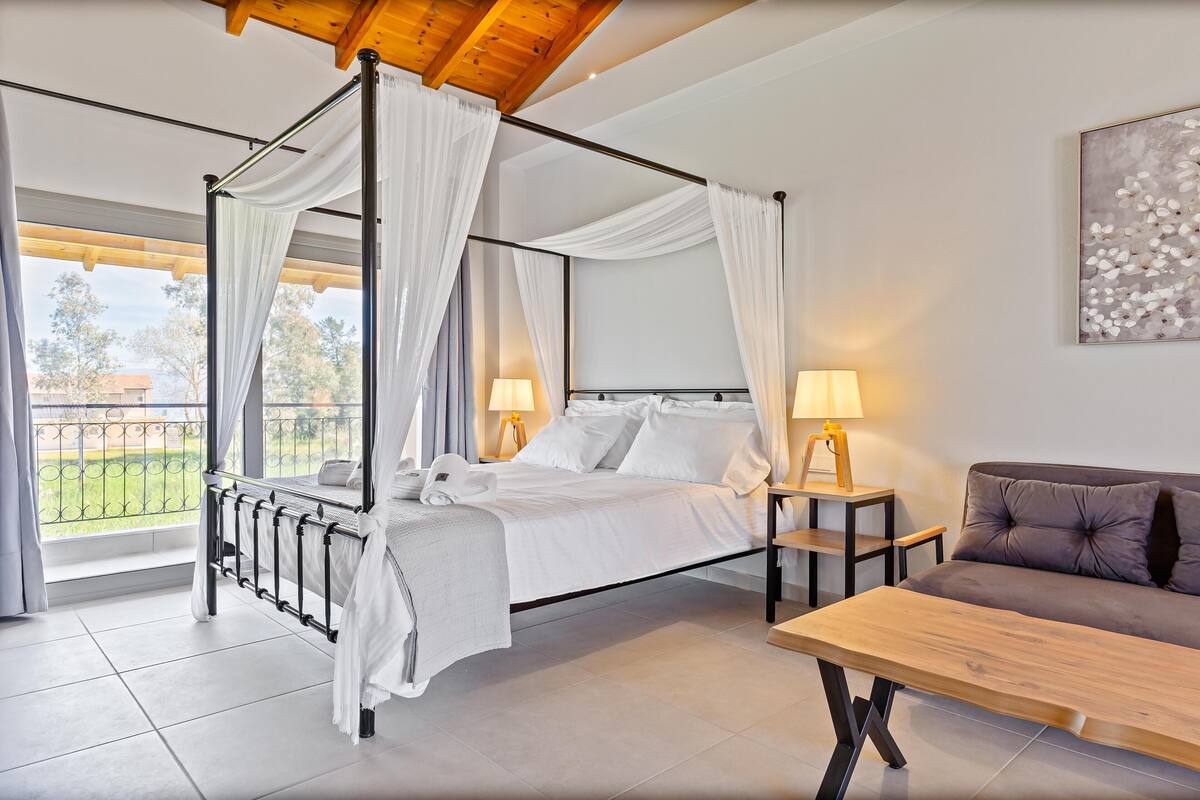 A four-poster canopy bed with white linens, two bedside lamps, a wooden table, and a sofa.