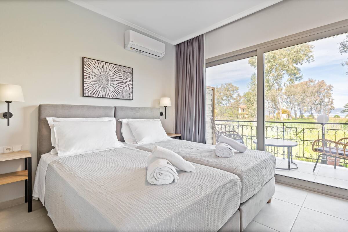 A hotel room with two beds, a balcony with a table and chairs, and a large window offering a view of trees and a body of water.